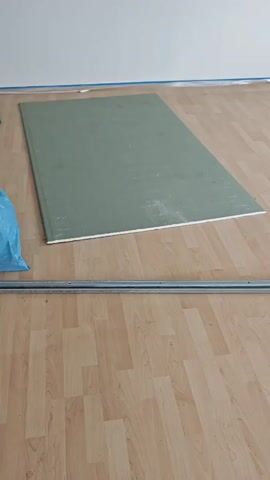 A single sheet of green drywall lies flat on a light-colored wooden floor. To the left of the drywall, a blue plastic bag is visible. In the foreground, a metal track runs horizontally across the scene. In the background, the bottom of a white wall is present. The lighting suggests an indoor environment.