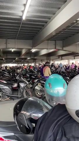 The image depicts a bustling motorcycle parking area, likely at an indoor market or event in Wirosari, Indonesia. The scene is filled with a vast number of motorcycles parked closely together, suggesting high demand for transportation or a popular gathering. Several people, predominantly women wearing headscarves, are visible navigating through the crowded parking lot or preparing to depart. The atmosphere appears busy and active, with the presence of numerous vehicles and individuals.

The setting is an indoor, covered parking structure with a corrugated metal roof supported by industrial beams. Fluorescent lights are visible, indicating it is likely daytime, though the enclosed space creates a consistent lighting environment. The motorcycles are predominantly dark-colored, with some featuring accents of red on their wheels. In the foreground, three helmets are prominent: a black full-face helmet, a bright teal helmet, and a white helmet, suggesting that some individuals have just arrived or are about to leave.

The activity taking place is the movement and management of motorcycles within a packed parking area. The expressions of the people are not clearly discernible due to the distance and resolution, but the density of the crowd implies a lively and possibly hurried environment. There are no immediately obvious signs of distress or negative emotions, rather a sense of everyday activity. The location cues, such as the style of motorcycles and the attire of the people, are consistent with an Indonesian context. The overall impression is one of a busy day at a popular spot in Wirosari.