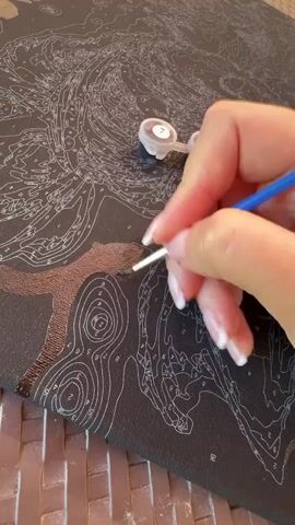 A person with manicured fingernails paints a dark canvas with a thin brush, filling in numbered sections with brown paint. A small paint pot, labeled "7," rests on the canvas near their hand. The canvas has white lines and numbers marking out a complex pattern. The person is wearing a light-colored shirt or top.