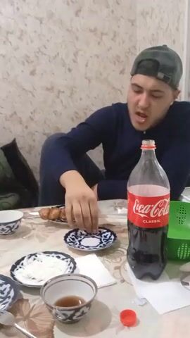 The video shows two men sitting at a table eating. The table is covered with a patterned tablecloth and set with plates, bowls, and drinks. The first man, wearing a dark blue sweater and a green cap, is shown eating from a skewer of grilled meat. There is a bottle of Coca-Cola on the table in front of him. The camera pans to the second man, who is wearing a black shirt. He is holding a cigarette and is also eating. A teapot, plates of food, and a phone are on the table near him. The walls of the room have a light-colored, textured pattern. A green jacket is visible on the back of a chair behind the table.