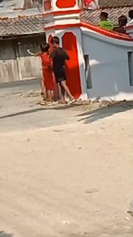 The video shows two young boys standing near a red and white structure, possibly an archway or gate, in an outdoor setting. The boy on the left is wearing a red shirt with the number 8 on the back and dark shorts. The boy on the right is wearing a black t-shirt and black shorts. They appear to be interacting, though their specific actions are unclear due to the video quality. Behind them, there is a building with a tiled roof and another structure decorated with red and white. A motorcycle is visible passing by in the background, and several other people are seen in the distance, some standing on the structure. The ground is unpaved and appears to be dirt or sand. The overall environment suggests a rural or semi-rural area.