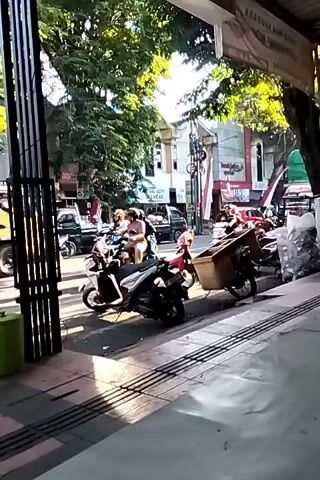 The video shows a street scene in what appears to be Southeast Asia. Several motorcycles are parked along the sidewalk, with some riders seated on them. A truck with a green tarp covering its load is visible in the background. There are trees lining the street and buildings on either side. The lighting suggests it is daytime, possibly late afternoon given the angle of the sun. The camera is positioned at a low angle, looking across the street from a sidewalk. The overall impression is a busy urban environment with a mix of vehicles and pedestrians.