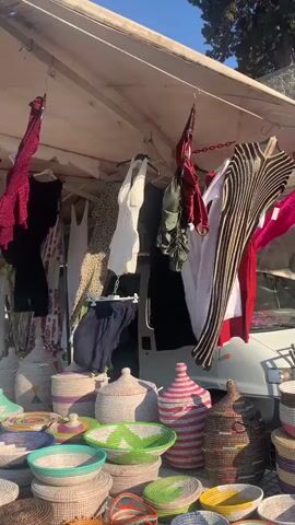 A market stall displays various items for sale. Hanging from a white awning are several pieces of clothing including dresses, swimsuits, and tops. In front of the clothing, a collection of woven baskets in various sizes and colors are arranged on a white tablecloth. The baskets are decorated with colorful stripes and patterns. In the background, a white van is partially visible. The scene is outdoors, with a clear blue sky overhead, suggesting a sunny day.