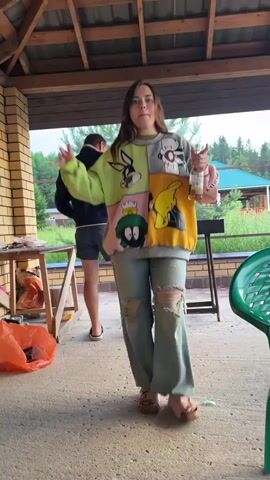 A young woman with brown hair, wearing a colorful Looney Tunes sweatshirt and ripped jeans, dances outdoors on a concrete patio. She holds a bottle in her right hand and makes hand gestures with both hands. A man is partially visible in the background, wearing a dark t-shirt and shorts. The scene is set under a covered patio with a wooden roof. In the background, trees and a house are visible. There are various objects scattered around, including a green plastic chair, a table with items on it, and an orange trash bag. The lighting suggests it is daytime.