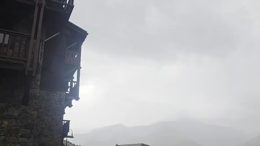 A wide shot of a mountain village on a cloudy day. On the left is the corner of a stone and wood building with balconies. In the center and right are several wooden chalets with dark roofs. In the foreground on the left is the rear of a black car parked on a wet road. In the background, mountains are visible through the mist. A lamppost stands on the right side of the frame.