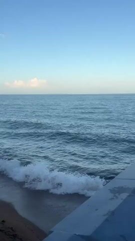 The video depicts a tranquil ocean scene with a clear blue sky and gentle waves lapping against a concrete barrier. The camera gradually pans from left to right, revealing a small island in the distance. The barrier has a decorative light fixture on top. The overall atmosphere is calm and serene.