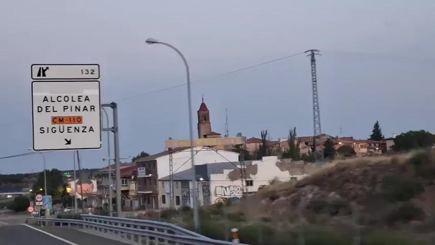 The video begins with a close-up of a road sign indicating "Alcolea Del Pinar CM-110 Siguenza" with an arrow pointing right, and a number "132" on the upper right corner. The sign is mounted on a metal pole. In the background, a town is visible with buildings, some of which appear aged and have graffiti. A prominent church tower with a red roof stands out against the dusky sky. The camera then pans slightly to the right, revealing more of the town's architecture, including residential buildings and some commercial structures. There are also several road signs visible on the left side of the frame, including speed limit signs (60 and 50). The environment appears to be rural or semi-urban, with trees and vegetation also present. The sky is overcast and suggests either early morning or late evening. There are no people clearly visible in the frame, and the primary focus is on the road sign and the townscape.