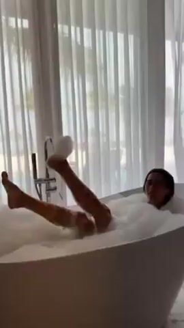 A woman is lying in a large, white, freestanding bathtub filled with bubbles. She is facing the camera and smiling. Her legs are extended, and her feet are above the water line. The bathroom has a large window with white sheer curtains, and palm trees and a beach are visible outside. A modern chrome faucet stands beside the tub.