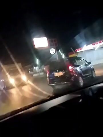 The video, shot from inside a vehicle at night, captures a street scene with several vehicles and a gas station. In the foreground, the dashboard and windshield of the recording vehicle are visible. Through the windshield, a gas station with red and yellow signage is illuminated by bright lights. Several cars and trucks are seen on the road. One car with purple taillights is in the middle ground, and a white truck with bright headlights is in the left midground. Further down the road, another truck, possibly a flatbed carrying a large cargo container, is visible. The road appears to be paved, and there are streetlights and building lights in the background. The overall impression is a nighttime view of a busy road with commercial establishments.