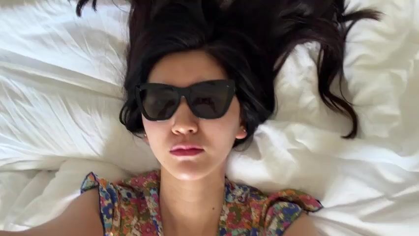 A woman with long dark hair and wearing black sunglasses is lying on her back on a white bed. Her hair is spread out around her head. She is wearing a colorful floral-patterned top. The lighting is bright. There are no visible actions or interactions.