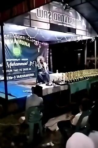 The video shows a man speaking from a stage in an outdoor tent setting. There are rows of trophies on a table to his right. He is speaking to an audience seated on plastic chairs. A backdrop includes religious text. Shoe racks are visible behind the audience. The lighting is dim.