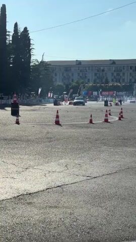 A dark colored car is drifting around an asphalt track on a sunny day. There are traffic cones placed around the track. In the background, there is a large building with banners and people watching the event.