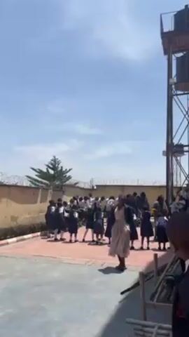 On a sunny day, a group of children in school uniforms stand in a courtyard, potentially a schoolyard or similar outdoor space. A woman in a patterned dress stands in front of them, possibly instructing or leading them. Behind the children is a wall topped with barbed wire, and to the right, a large water tower stands tall. The setting suggests a school environment, possibly in an area with visible security measures. The children appear to be engaged in an activity, likely organized by the woman in front of them. The sky is bright blue with scattered clouds. The ground is paved, and there are some wooden structures visible at the bottom right corner of the frame.