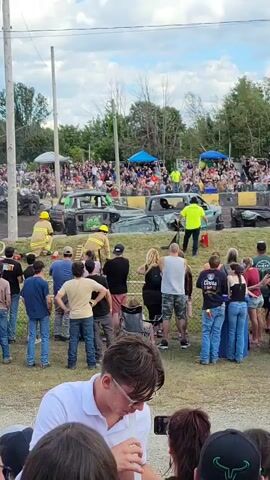 A demolition derby event is underway. Several cars have crashed and are smoking. Firefighters in yellow helmets and suits are on the scene attending to the vehicles. A crowd of spectators is gathered behind a fence watching the event. The environment appears to be an outdoor arena with a dirt track. The sky is partly cloudy.