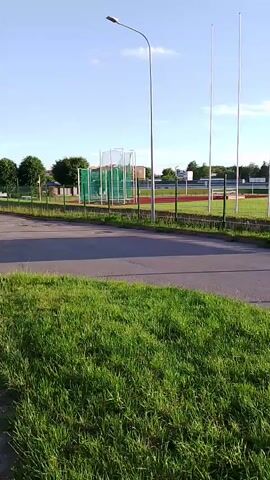The video pans across an outdoor sports field, likely a track and field stadium. In the foreground, a patch of green grass lines a paved surface, possibly a road or pathway. Beyond this, a black metal fence encloses the field. The field itself appears to be a track with red lanes and a central grassy area. Several tall, slender white poles, possibly for high jump or flags, are visible on the field. To the left, a green netted structure, possibly for discus or hammer throw, is present. Further in the background, buildings and trees are visible under a clear blue sky with scattered clouds. A large, blank billboard structure stands to the right of the frame, supported by metal scaffolding. The scene is brightly lit by sunlight, casting shadows on the paved area. There are no people visible in the video.