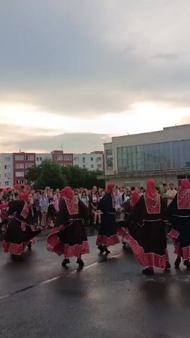 A group of five women in traditional folk costumes are performing a synchronized dance outdoors in front of an audience. The women are wearing dark dresses with red and white patterned trim on the skirts and headscarves. The sky is cloudy and appears to be late afternoon or evening. Buildings, including apartment blocks and a larger modern building, are visible in the background. The ground is wet, suggesting it has recently rained. The audience is gathered behind a barrier, watching the performance.