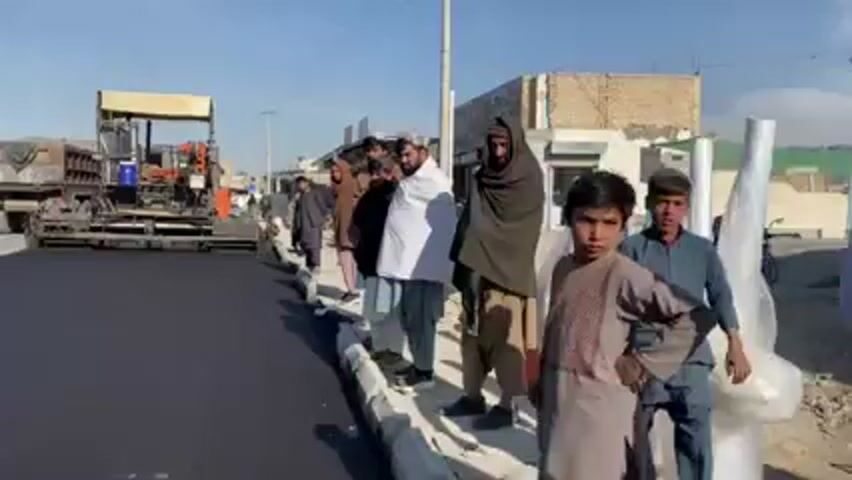 The video shows a group of people standing on the side of a newly paved road. The road is black and smooth, and a large paving machine is visible in the background. The people are dressed in casual clothes, some in traditional attire, and appear to be observing the road paving process. There are buildings in the background, suggesting an urban or semi-urban setting. The sky is clear and blue, indicating a sunny day.