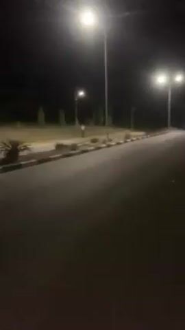 The video shows a dark road at night, illuminated by streetlights. On the side of the road, there is a grassy area with small plants. In the middle of the video, a person is seen walking along the side of the road.