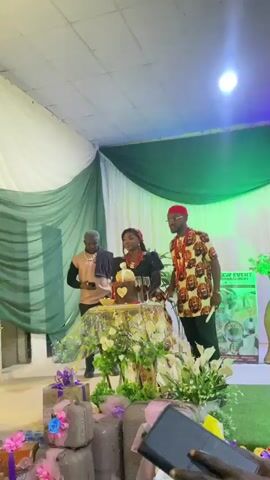 The scene showcases three individuals standing behind a table adorned with flowers and a cake, likely at a wedding or celebration. The woman in the center, presumed to be the bride, is dressed in a black and red traditional outfit. A man to her right is wearing a vibrant patterned shirt and a red cap. To her left, another man is in a brown top and a cap. They are standing on a slightly elevated platform that appears to be a stage, decorated with white and green drapes. There are decorations, including wrapped gifts, around the base of the table. The room is lit with a blue hue, and other attendees are visible in the foreground, holding up their phones to capture the moment. The background suggests an event venue or hall.