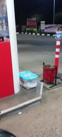 The video begins with a close-up of a red and white structure, likely a gas pump at a gas station at night. A blue and white box is visible on the ground next to it, along with a red trash can. The camera then pans to the right, showing more of the gas station equipment and a blurry background of a road with vehicles. The scene transitions to focus on a person wearing a red hooded jacket, standing next to a motorcycle. The person appears to be interacting with the motorcycle's storage compartment, which is open. They are holding cash in their hands and looking at the camera. The person then gestures with their hand and appears to be putting the cash into the motorcycle's storage. The video continues to show the person at the gas station, with the motorcycle and gas pump in the foreground. The person is seen holding and looking at the money. The ambient lighting suggests it is nighttime. The overall scene depicts a transaction or interaction involving money at a gas station.