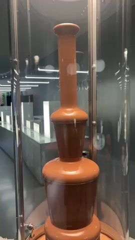 The video shows a chocolate fountain. The fountain is enclosed in a clear cylindrical container, revealing the chocolate flowing from tier to tier. The base of the fountain is stainless steel. A woman's hands are shown using a white plastic spoon to catch chocolate from a spigot at the base of the fountain. The text on the stainless steel base reads, "BITTE ZIEHEN, PLEASE PULL", followed by information about allergen content in German and English. The background reveals a modern, brightly lit space with white countertops. The camera moves slightly as the chocolate is being sampled.
