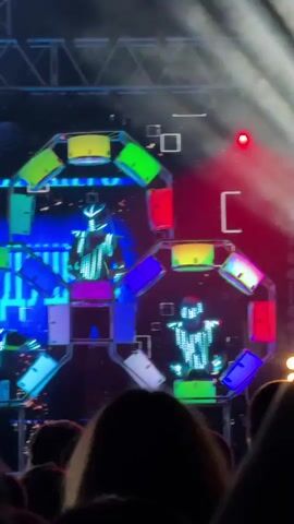 A live performance stage is seen with multiple performers in illuminated costumes. The stage features a large, abstract structure made of panels, many of which are lit with different colors, including blue, purple, green, red, and white. The performers are positioned within and around this structure. In the center, a performer stands on a raised platform wearing a suit that flashes with white lights. To the left and right, two other performers in similar illuminated suits are visible. The background is dark, with some bright blue lights and what appear to be small, floating white particles. A disco ball is visible in the upper left corner. The lower part of the frame shows the silhouettes of a crowd of people watching the performance. The overall atmosphere is that of a high-energy, visually dynamic live show.