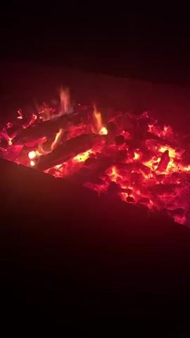 This media file shows a close-up view of burning embers and logs in a fire pit. The dominant colors are vibrant reds and oranges, indicating intense heat. Small flames dance among the glowing coals, with wisps of blue and white visible in places. The scene is dark around the edges, suggesting it is nighttime or the fire pit is in a shaded area. The focus is entirely on the fire, creating a sense of warmth and intensity. There are no people visible, and no discernible text. The setting appears to be an outdoor fire pit, possibly in a backyard or designated fire area. Given the location context of Taganrog, Russia, this could be a common scene during cooler evenings or for outdoor gatherings. The mood conveyed is one of primal energy and comfort associated with a campfire.