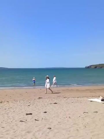 The video displays a beach scene under a clear blue sky with scattered clouds. A calm ocean stretches towards the horizon, meeting a coastline with green hills and some buildings. Several people are present on the sandy beach and in the shallow water. On the beach, some individuals are sunbathing, while others walk along the shoreline or in the water. In the foreground, a person is seated on a towel, facing away from the camera. Further down the beach, a few more people are visible, including a couple walking and another person standing near a beach chair. In the distance, more people are scattered across the beach and in the sea, suggesting it is a moderately busy day. The overall atmosphere appears relaxed and summery.