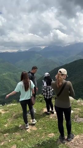 The video begins with a shot of four young adults, two males and two females, standing on a grassy cliff edge overlooking a vast, verdant mountain range under a cloudy sky. One woman is facing away from the camera, wearing a light blue shirt and black pants, while a man stands next to her, looking towards the mountains. Another woman, in a black and white plaid shirt and black pants, is looking at her phone. The fourth person, a woman wearing a green long-sleeved shirt and black pants, has her back to the camera and appears to be adjusting something on her shoulder.

The camera then pans to the right, revealing another woman sitting on a rock formation on the same cliff, wearing a light grey tracksuit and sunglasses, with a mountainous backdrop. Further to the right, another person is visible sitting on the edge of the cliff.

The perspective shifts again, showing the woman in the green shirt from the first shot adjusting her hair, with the mountainous landscape behind her. The camera then returns to the group of four, who are now closer together, with the woman in the plaid shirt showing something on her phone to the man.

The video concludes with a sweeping panorama of the extensive mountain range, showcasing layers of green, forested hills receding into the distance, with snow-capped peaks visible on the horizon under a dramatic, cloud-filled sky. A winding road is visible in the valley below. The foreground shows a grassy slope with rocks.