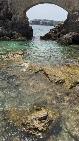 The video shows a view of the ocean through a stone archway. The water is clear and turquoise, with rocks visible beneath the surface. In the distance, there are buildings and a body of water. The sun is shining, casting a bright light on the scene.