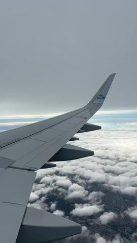 The video shows an aerial view from an airplane. The right wing of the plane is visible in the foreground, with the KLM logo on the wingtip. Below the wing, there are layers of white clouds with glimpses of a city or land below. The sky above is overcast with gray clouds. Text overlays indicate that the gate will close in 15 minutes before departure.