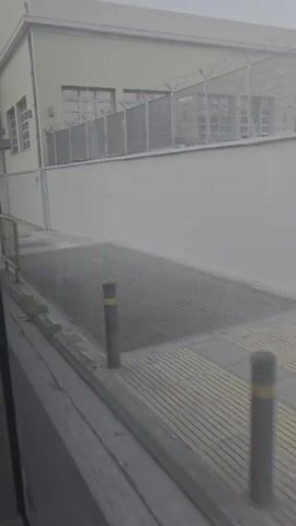 The video shows a street scene passing by from a vehicle. On the left, there is a light-colored building with windows and a fence on top. A sidewalk with a yellow railing is visible. The ground is paved, and there are some trees. A street sign with a red and blue circular symbol is also present. The weather appears to be overcast or hazy.