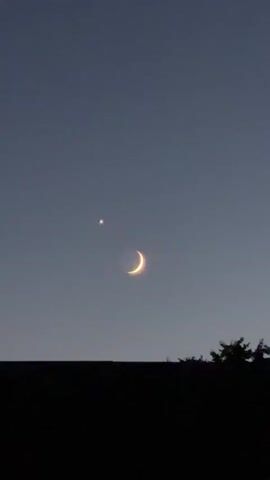 The video shows a dark silhouette of a roofline in the foreground with a dark silhouette of a tree on the right side. The sky above is a deep twilight blue. In the sky, a crescent moon is visible, glowing with a bright yellowish-orange light. To the left of the moon, a small, bright, star-like object, likely a planet, is visible. The moon and the star-like object appear to be close together in the sky. There are no people or other discernible objects or actions in the video. The scene appears to be a still image captured at dusk or dawn.