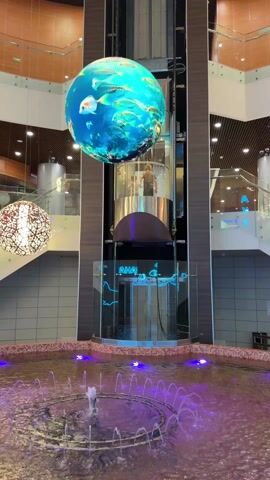 The interior of a modern shopping mall is displayed. At the center is a round fountain illuminated with purple lights, with small jets of water spraying upwards. Above the fountain is a cylindrical glass elevator, with people inside moving upwards. Beside the elevator is a large spherical LED display showing a scene of fish swimming underwater. The mall has multiple levels, with glass railings lining the staircases. Several decorative light fixtures hang from the ceiling. Two individuals are standing on a higher level of the mall, conversing. The color palette consists of blues, browns, and whites, creating a clean and contemporary ambiance.