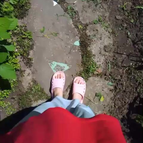 The video shows a first-person perspective of someone walking on a cracked stone path outdoors. The person is wearing light blue pants and pink slides. The path is surrounded by greenery and dirt. There are shadows cast on the path from what appears to be overhead foliage. The person's red top is visible in the foreground, obscuring the view of their torso. A green watering can is visible to the left of the path in the earlier frames. The scene appears to be a garden or yard.