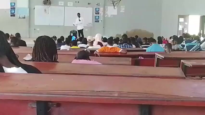 A wide-angle shot shows a classroom filled with students sitting at wooden desks facing a person standing at the front. The speaker is wearing a white shirt and dark pants and appears to be addressing the class. The desks are arranged in rows, and the students are a mix of genders and ethnicities. Some are looking at the speaker, while others appear to be looking down or talking to each other. There are whiteboards and posters on the walls. The lighting in the classroom is bright.