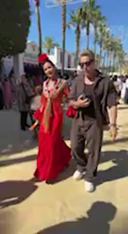 This video captures a lively outdoor scene, likely a fair or festival, in Jerez de la Frontera, Spain. The main subjects are a couple walking arm-in-arm. The woman, on the left, is wearing a vibrant, traditional red flamenco dress with a full, ruffled skirt and a matching red flower adorning her hair. She also has an intricately embroidered red and gold fringed shawl (mantón de Manila style) draped over her shoulders. She smiles and briefly turns to face the camera before continuing her walk. The man, on her right, is dressed in a dark short-sleeved shirt, dark pants, and sunglasses.

The setting is bright and sunny under a clear blue sky. Palm trees are visible in the background, along with numerous other people and various temporary structures, some featuring red awnings, suggesting a bustling public event. The ground appears to be a light-colored, possibly paved or dirt, area. The traditional attire of the woman strongly indicates a cultural celebration, such as the famous Feria del Caballo (Horse Fair) which takes place in Jerez and is known for its elaborate flamenco fashion and festive atmosphere. The couple appears joyful and relaxed, enjoying the festive occasion. There is no discernible text visible in the video.