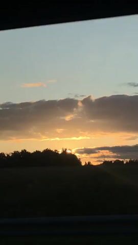 A time-lapse video captures the sunset over a rural landscape. The sun, a bright yellow orb, is partially obscured by a silhouetted line of trees and a low hill. Wispy clouds, tinged with orange and pink, stretch across the sky, which transitions from a pale blue at the top to a warmer yellow near the horizon. In the foreground, a dark, blurry band suggests the presence of a road or a vehicle's interior, with a faint metallic railing visible at the bottom. The overall impression is of a serene evening as daylight fades.