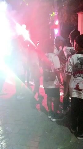 A group of people are gathered at night outdoors. One person in the center is holding a flare that emits red smoke and light, illuminating the surrounding area and the faces of the people. Other people are visible in the background, some holding flags or banners, and there appears to be some form of stage or lighting setup in the distance. The overall atmosphere suggests a celebration or event.