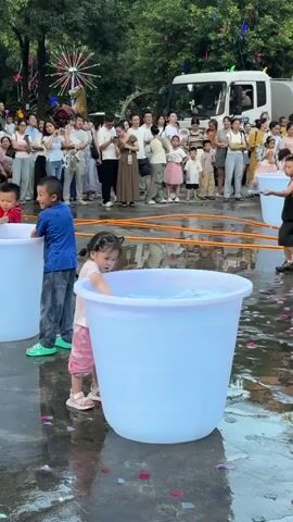 A group of children are gathered around large blue tubs of water in what appears to be an outdoor event, possibly a festival. Several children are actively engaged in playing with the water. In the foreground, a young girl in pink shorts and a white t-shirt is reaching into a tub. To her left, a boy in a blue shirt and jeans is also near a tub, with his hand in the water. Further to the right, another boy in a black tank top and shorts is holding a long, thin tool, possibly a water gun or scoop, and appears to be aiming it or interacting with a tub. Behind the children, a crowd of adults and more children are observing the activity. They are standing behind orange hoses that are laid out on the wet ground. In the background, a white truck with some writing on its side is visible, and there are decorative lights and foliage suggesting a celebratory atmosphere. The ground is wet and littered with colorful pieces of paper or petals. The scene is brightly lit, likely during the daytime.