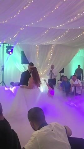 A wedding reception with a couple dancing in the center of the frame, surrounded by artificial fog and purple and green lighting. The bride wears a white gown and the groom a dark suit. Guests are seated at tables in the foreground and background, with some looking towards the dancing couple. A man with a microphone stands in the background. The ceiling is draped with white fabric and string lights.