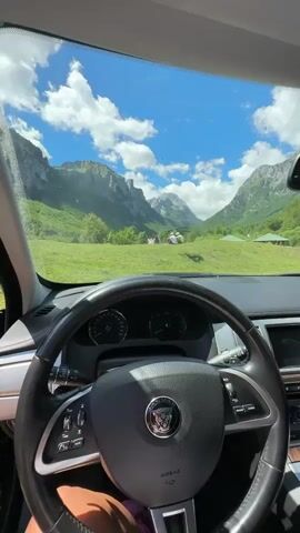 The video begins inside a car, showing the steering wheel and dashboard. The view through the windshield reveals a picturesque mountainous landscape with lush green fields and a clear blue sky dotted with white clouds. The camera then pans slightly to the right, revealing the open car door and a broader view of the scenic outdoor environment. The scene transitions to focus on the expansive green meadow with several people sitting on the grass, enjoying the view of the majestic mountains in the background.
