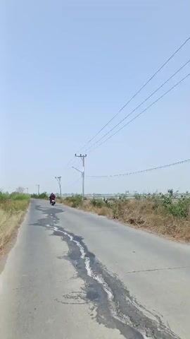 A motorcycle is traveling down a paved road under a clear sky. The road has patches of damage and discoloration. On the left side of the road, there are tall grasses. On the right side of the road, there are bushes, dried grass, and a body of water. Telephone poles with power lines are visible along the road. The motorcycle appears to be red and is moving away from the camera.