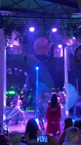 The video shows a group of performers on a stage in what appears to be a restaurant or club. The performers are wearing costumes, and some are holding large fans. The stage is decorated with plants and lights, creating a festive atmosphere. There are people in the audience watching the performance, and some are holding up their phones to record it. The lighting is predominantly blue and green, with spotlights shining on the performers.