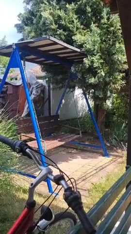 The video shows a backyard scene with a swing set and a trampoline. A bicycle is partially visible in the foreground. The camera pans to the right, revealing a wooden structure, possibly a gazebo, with a table and chairs. Lush green trees and bushes surround the area, suggesting a warm, sunny day. The overall impression is that of a recreational space in a home garden.
