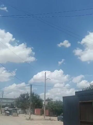 This media file captures an outdoor scene on a bright, sunny day in Zhanaozen, Kazakhstan. The sky is a vibrant blue, dotted with fluffy white cumulus clouds. Overhead, several power lines crisscross the frame, adding a sense of utility to the otherwise natural sky.

The foreground features a partially visible dark-colored, modern-looking building with large windows, suggesting it might be a commercial establishment or an office. To the left, a street or dirt road is visible, with a few indistinct figures walking in the distance. There are also utility poles, one of which has a security camera mounted on it. In the middle ground, trees with green foliage are interspersed with what appear to be shipping containers, one of which is red. A silver car is parked on the right side of the road.

The overall atmosphere is one of a typical day in a town or city, with a mix of modern infrastructure and natural elements. The bright sunlight indicates it is likely midday, and the clear skies suggest fair weather. There is no discernible text within the frame. The scene appears calm and ordinary, without any specific event unfolding, but rather a snapshot of daily life in this location.