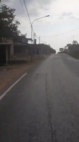 The video shows a POV perspective of someone walking on a paved road that slopes upwards. The road is cracked and has patches. On the left side of the road, there is a dry grassy area with some buildings and structures. On the right side, there are more structures and some trees, including palm trees. Overhead, there are utility wires and poles. The sky is cloudy with some patches of brighter light suggesting the sun might be attempting to break through. The overall environment appears to be a rural or semi-urban area with a slightly overcast sky.