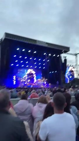The video shows a large outdoor concert stage with a banner reading "SUMMER SESSIONS." The stage is lit with blue and white lights, and there is a large screen displaying a silhouette of a person. A band is performing on stage, and a crowd of people is gathered in front of the stage, watching the performance. The sky is overcast. People are wearing casual clothing, and some are holding umbrellas, suggesting it might be raining or threatening to rain. The perspective is from the audience, looking towards the stage.
