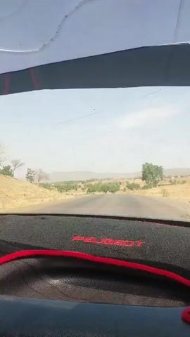 The video shows a view from inside a car driving down a paved road. The dashboard is visible, with a black cover and the word "PEUGEOT" stitched in red. The windshield has a few smudges and a crack.  The road stretches straight ahead through a dry, open landscape with scattered trees and low hills in the distance. The sky is pale blue. There are no other vehicles or people immediately visible in the video.