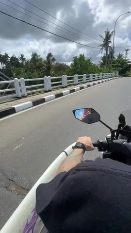This is a first-person perspective video from a motorcycle in Denuwala, Sri Lanka. The rider, wearing a black long-sleeved shirt and a smartwatch, is holding the handlebars of a scooter. A white surfboard is strapped to the side of the scooter. The rider is wearing a black helmet with stickers on it, one of which reads "SVOYDOSKU" and another has a "500" logo.

In the background, a second person wearing a helmet and a brightly colored floral top is visible on the back of the scooter. The weather appears to be sunny with scattered clouds, indicating daytime. The setting is a paved road lined with a white and black striped curb and a white railing. Lush green vegetation and palm trees are visible on either side of the road, suggesting a tropical environment. In the distance, there are buildings and utility poles. The mood seems to be relaxed and adventurous, as the individuals are cruising on a motorcycle with a surfboard. The overall impression is of a casual journey to or from a surfing spot in a scenic, tropical location.